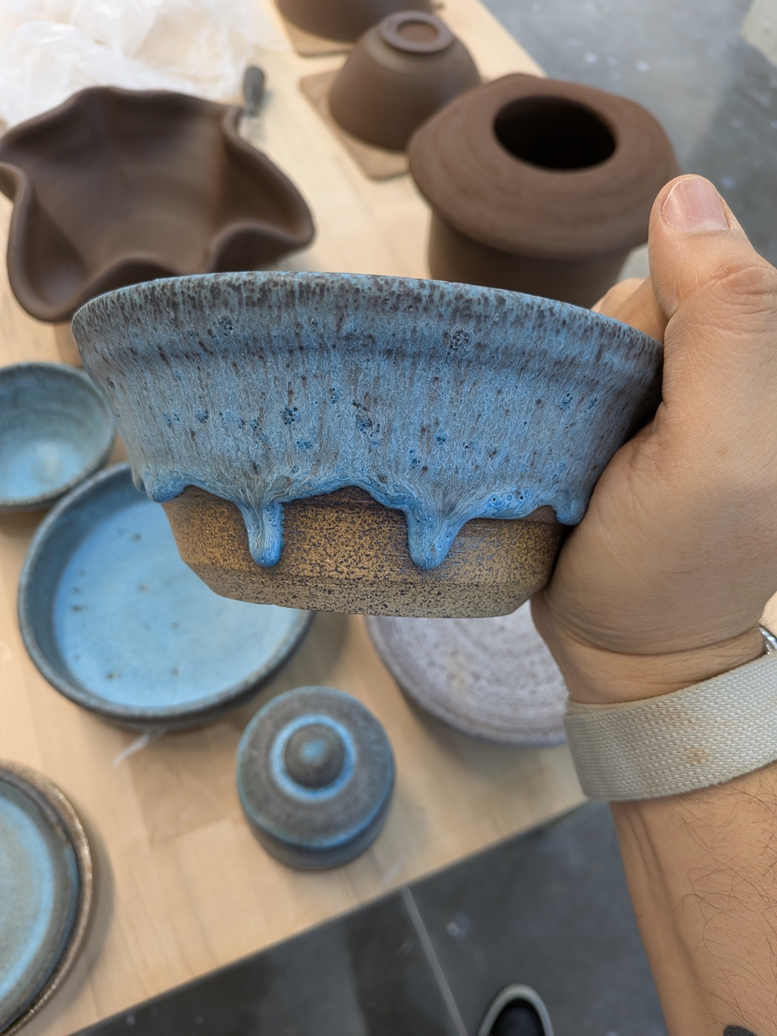 A glazed bowl held in the studio — blue glaze breaking over speckled stoneware, with unfired pieces on the table behind
